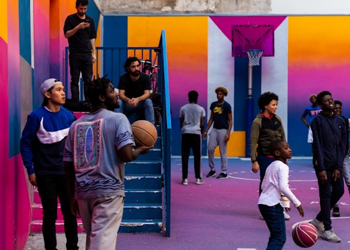 A vibrant outdoor basketball court filled with people of various ages engaged in different activities. Some are playing basketball while others are standing and chatting. The court is surrounded by brightly painted walls in shades of pink, orange, and blue. A basketball hoop is mounted on the back wall, adding to the sports theme. A few people are sitting on stairs or using phones, creating a relaxed and social atmosphere.