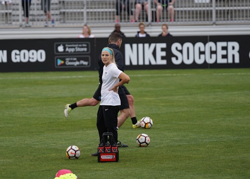 A person in sports attire stands on a soccer field, surrounded by several soccer balls and a carrier with bottles. The field is bordered by advertisements for Nike Soccer and app stores. Several people sit in bleachers in the background.
