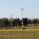 A sports field with a group of people engaging in athletic activities in the background. There is sports equipment like a soccer goal and a scoreboard visible. A chain-link fence runs across the foreground, and a statue or sculpture resembling an eagle is also seen. Trees and tall floodlights are positioned behind the field.