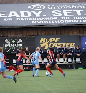 Players engage in a competitive soccer game on a field while others are seated on the bench watching attentively. The action is taking place in front of a wooden structure displaying multiple advertisements, including orthodontics, lacrosse, and field hockey.