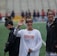 Several athletes in sports attire are on an outdoor sports field, interacting and smiling. One individual in the foreground waves with a friendly expression, while another stands beside her, also smiling. They wear protective goggles and gear associated with lacrosse. The background features indistinct figures in dark clothing against a backdrop of stadium seating.