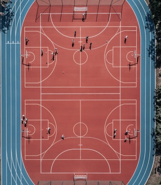 An aerial view of a large, vibrant sports complex featuring a red basketball court with multiple hoops and a surrounding blue running track. Several people are scattered across the area, engaged in various activities.