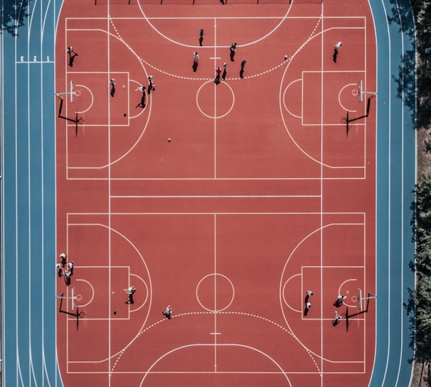 An aerial view of a large, vibrant sports complex featuring a red basketball court with multiple hoops and a surrounding blue running track. Several people are scattered across the area, engaged in various activities.