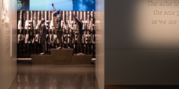 A dimly lit display features statues of athletes standing on a podium. They are positioned in front of a backdrop with the words 'LEVELING THE PLAYING FIELD' in bold letters. On the wall to the right, a softly lit quote is partially visible. The overall ambiance suggests a museum or exhibition setting.