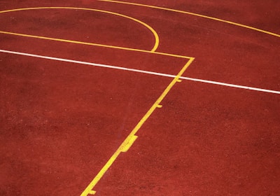 A sports court with a reddish-brown surface and bright yellow and white lines. The lines form geometric shapes, including rectangles and circles, suggesting markings for games like basketball or netball.