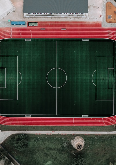 An aerial view of a large sports complex featuring a central green football field surrounded by a red running track. Adjacent to the track, there is a parking area, buildings, and pathways. Nearby, there are additional sports courts in different colors, including a blue swimming pool and orange basketball courts.