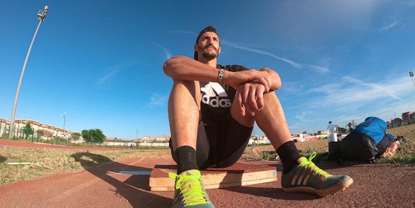 A person wearing a black athletic outfit is seated on a wooden platform in an outdoor sports field. The individual is relaxed, with arms resting on knees. The field is expansive with a clear blue sky and scattered clouds above. A brightly colored running shoe and athletic gear are visible, along with a water bottle and a blue backpack placed beside the person. Lampposts and residential buildings can be seen in the background.