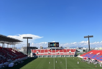 A large sports stadium with red and blue seating overlooks a green field with a goalpost at the center. A few people are scattered across the field, with some tents and a gathering of individuals on the right side. The sky is clear and blue with some clouds visible in the distance. A large screen or scoreboard is situated at the far end of the field.