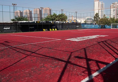 A sports court with a red playing surface and white lines, surrounded by a black mesh fence. In the background, tall buildings are visible, along with some trees and a few yellow chairs positioned near the fence.